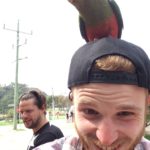 Giles Short selfie with parrot on head and friend in background