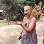 Blonde girl in dress excited with 1 parrot on her hand and one parrot on her head