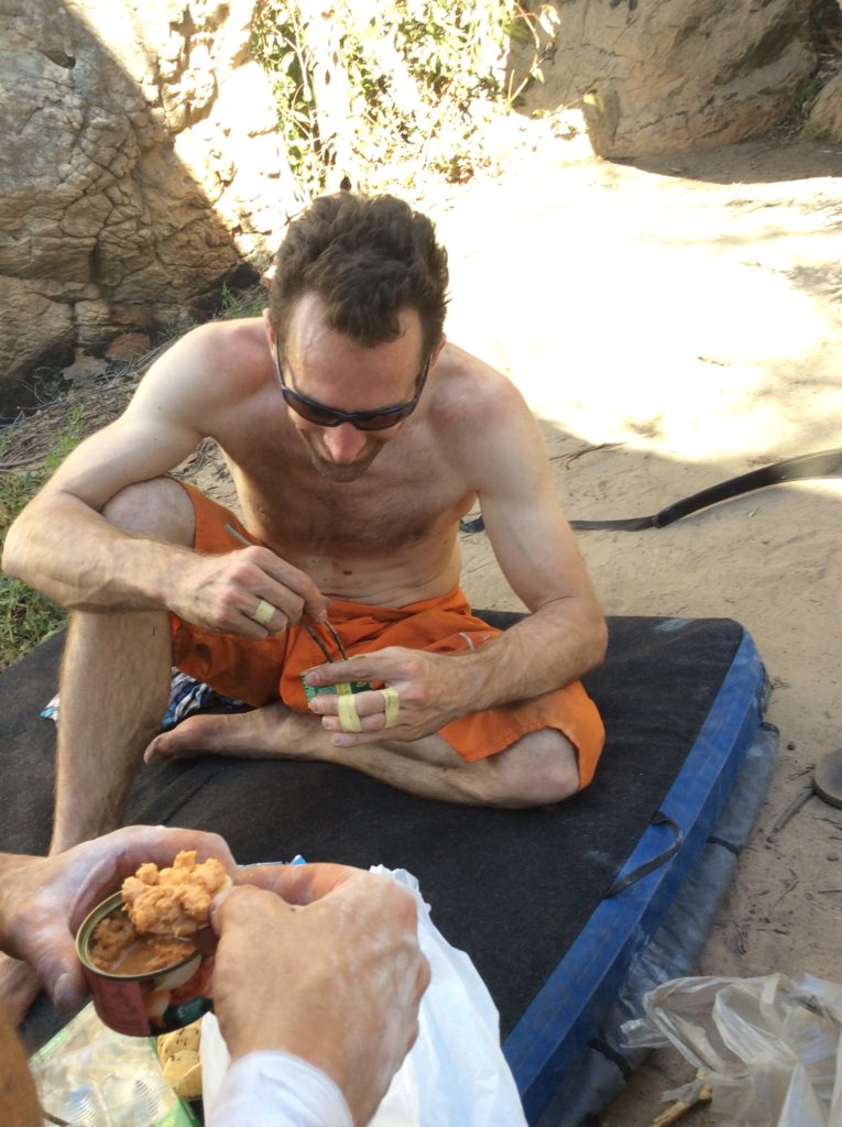 Vincent sat on bouldering mat using 2 sticks as chopsticks to eat can of tuna